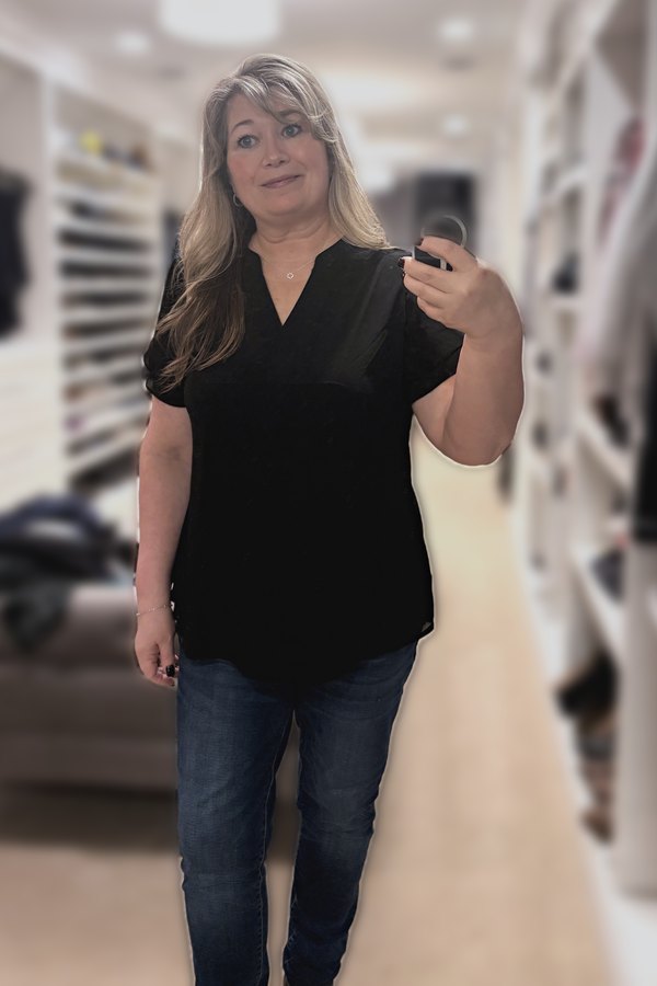 Mirror selfie of a woman wearing a black petal-sleeve V-neck top, dark jeggings, a dainty clover necklace and a silver bead bracelet